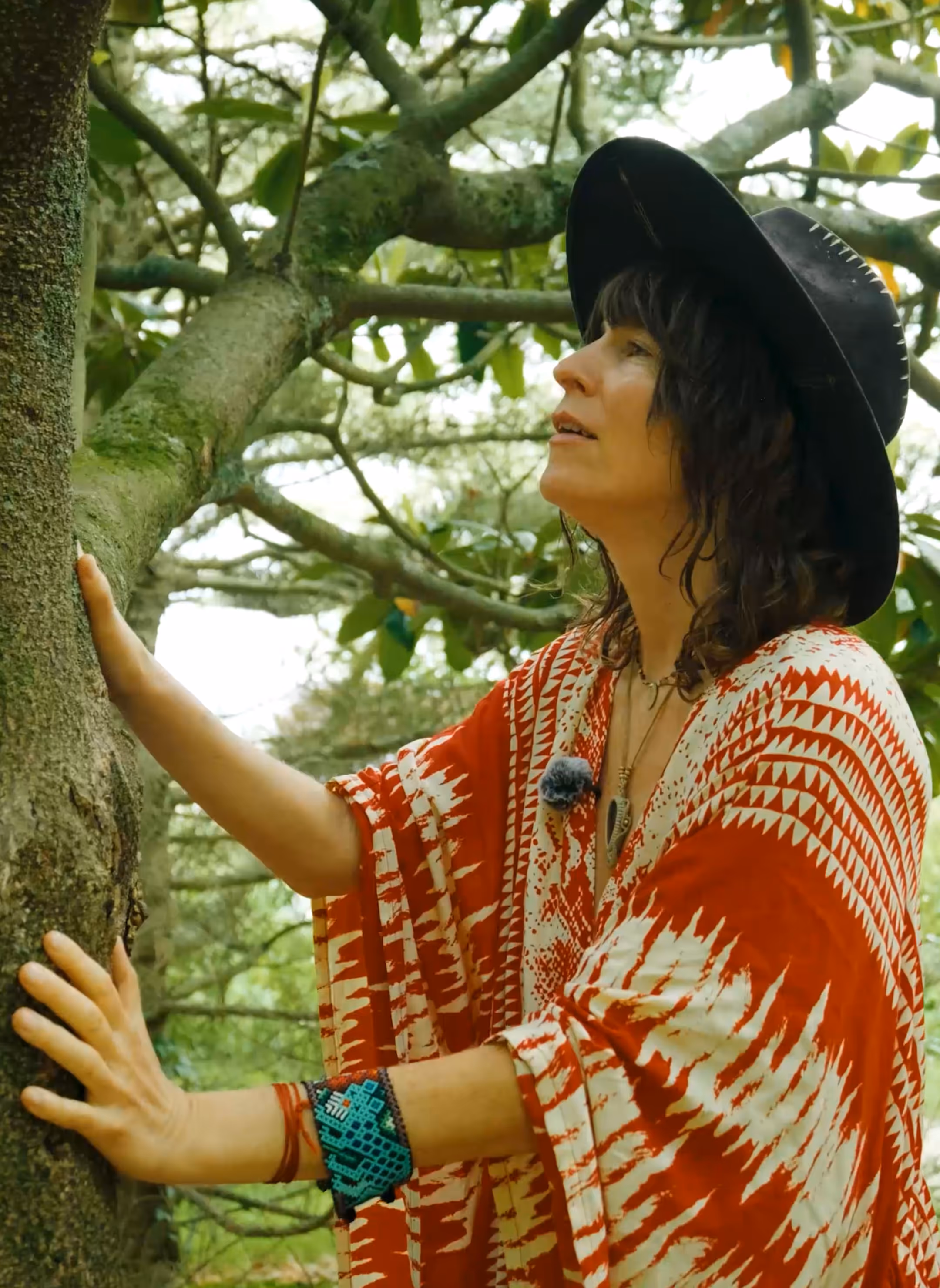 Person in black dress and wide-brimmed hat holding and playing a large frame drum while standing under a tree with shadows on a stone wall.