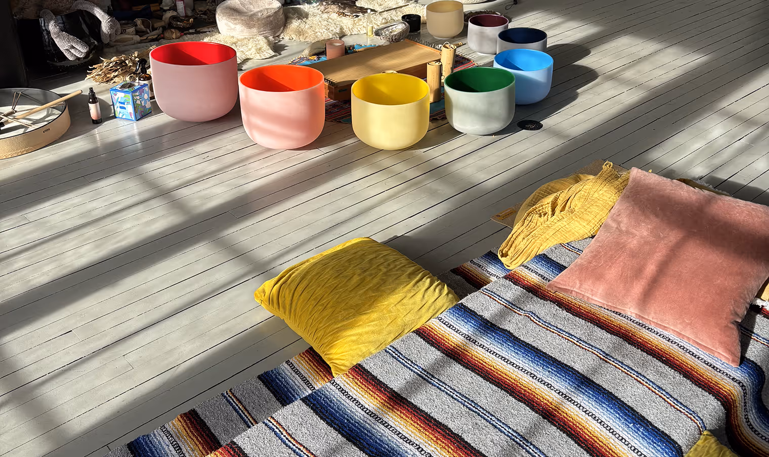 Sunlit room with colorful meditation cushions on a striped blanket and a set of vibrant crystal singing bowls arranged on a wooden floor.