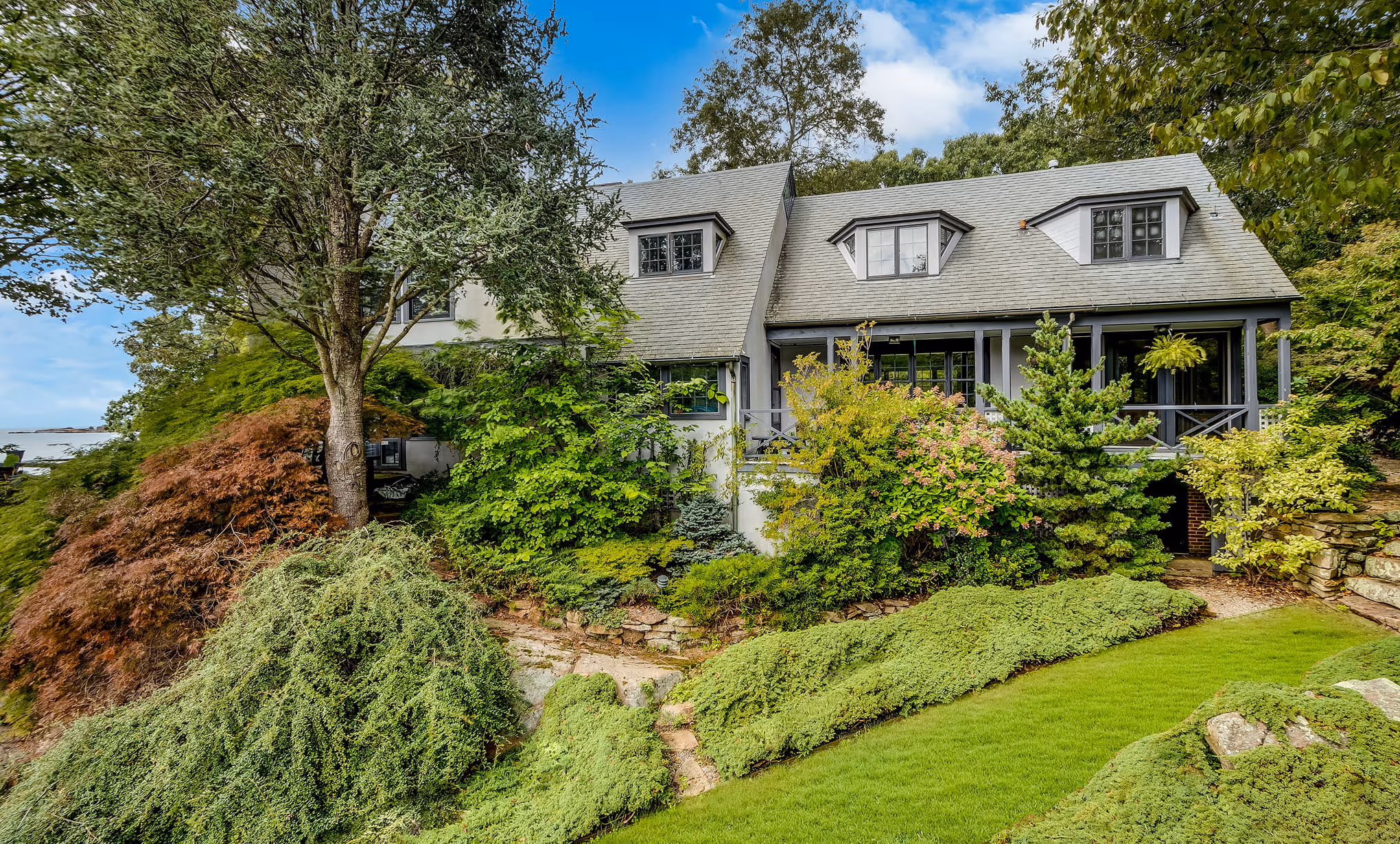 Gray house with dormer windows surrounded by dense green and autumn-colored shrubs under a partly cloudy sky.