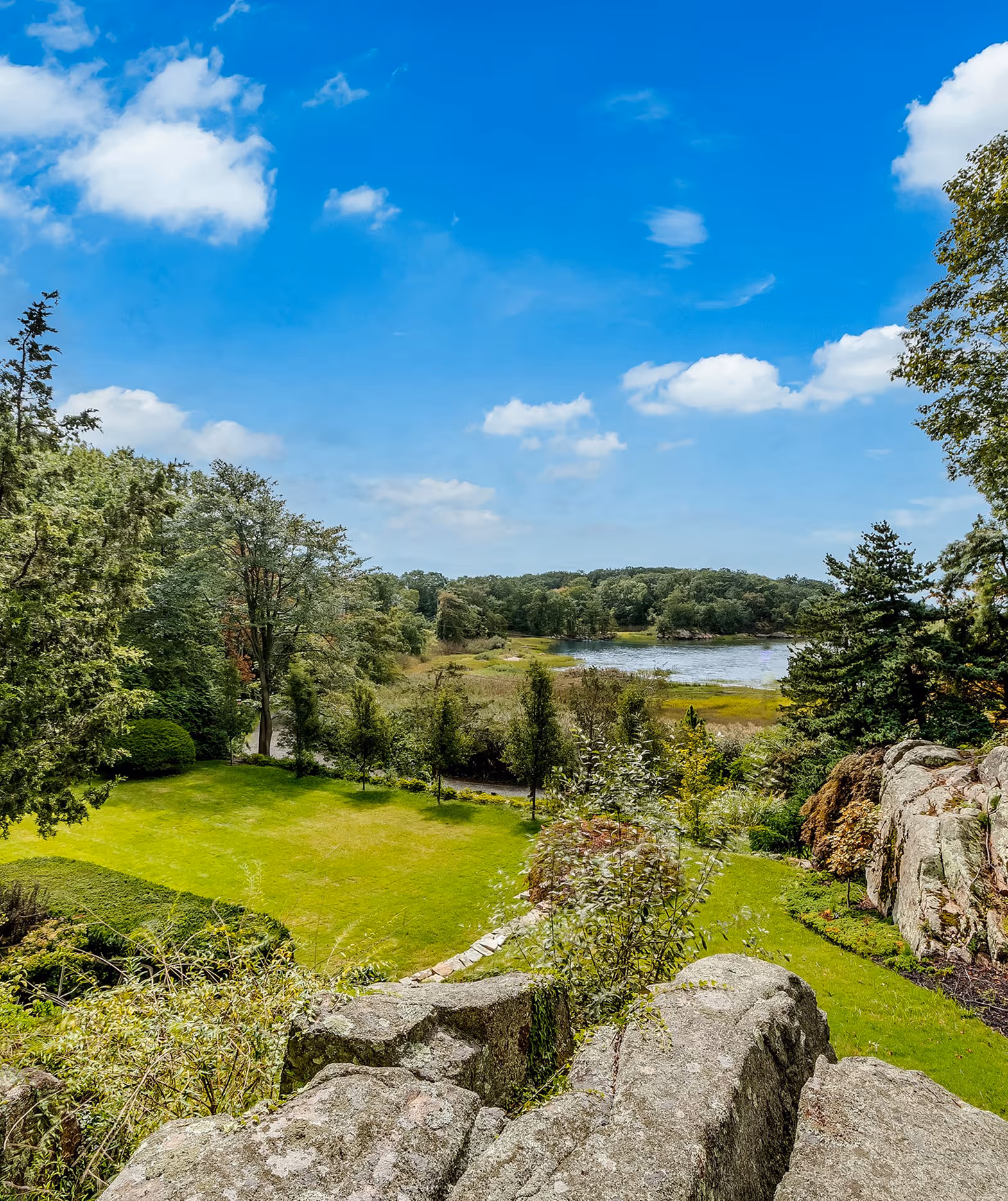 Green lawn and trees beside a body of water under a blue sky with scattered clouds.