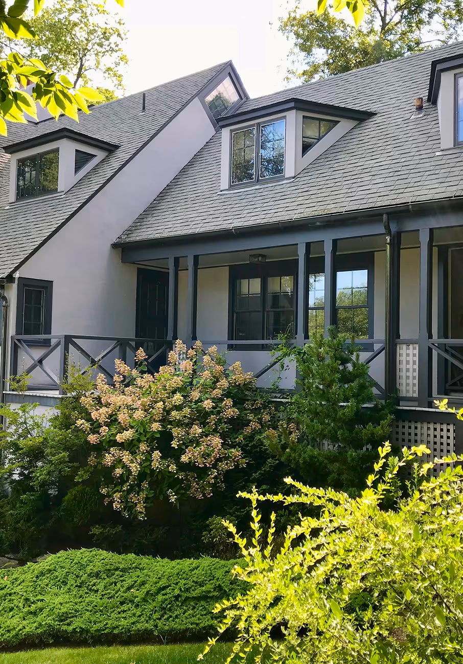 House with gray siding and dormer windows, a porch, and lush green shrubs in the front yard.