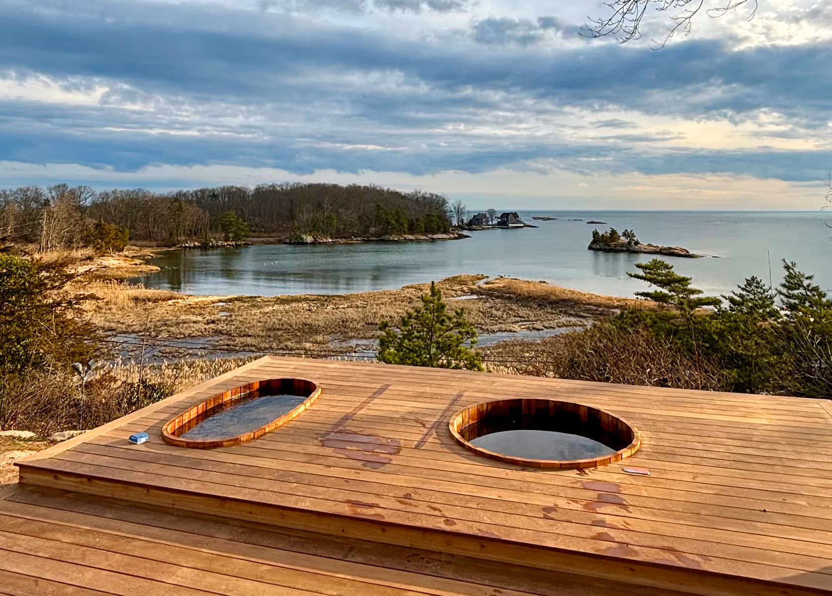 Wooden deck with two built-in hot tubs overlooking a calm coastal bay with trees and small islands.